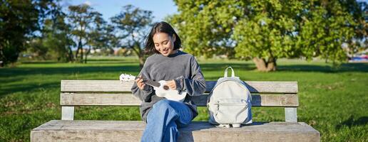mooi Aziatisch meisje Toneelstukken ukulele buitenshuis, zit in park Aan bank met musical instrument foto