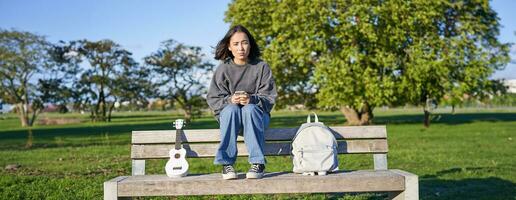 mooi brunette meisje Aan bank in park, zittend met ukulele en rugzak, Holding smartphone, gebruik makend van mobiel app foto