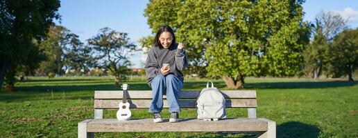 opgewonden meisje looks Bij smartphone en viert, wint Aan mobiel telefoon, zit met ukulele en rugzak in park Aan bank Aan zonnig dag foto