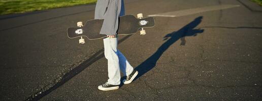 bijgesneden schot van tiener meisje lichaam, Holding kruiser longboard in hand, wandelen in sportschoenen Aan weg in jeans en trui. jong vrouw schaatser met skateboard foto