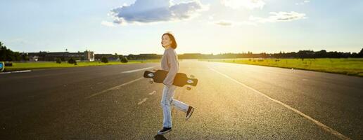 Aziatisch meisje met skateboard staand Aan weg gedurende zonsondergang. schaatser poseren met haar lang bord, kruiser dek gedurende opleiding foto