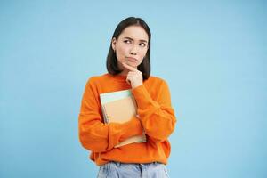 Aziatisch vrouw staand met notitieboekjes, denken, op zoek terzijde met attent gezicht uitdrukking, staand over- blauw achtergrond foto