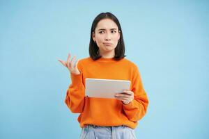 vrouw met digitaal tablet, looks verward, kan niet begrijpen iets Aan haar apparaatje, staat verbaasd tegen blauw achtergrond foto