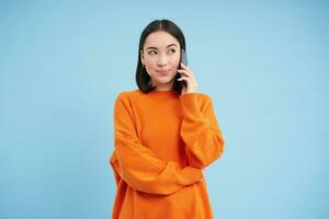 Koreaans meisje in oranje trui praat Aan mobiel telefoon, spreekt Aan mobiele telefoon met gelukkig glimlachen gezicht, bestellingen levering, overlegt met iemand Aan telefoon foto