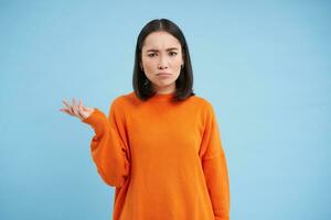 verbaasd Aziatisch vrouw haalt zijn schouders op, verhoogt een hand- en looks onwetend, kan niet begrijpen wat is gebeuren, staart met verward en boos gezicht, blauw achtergrond foto