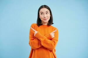 beslissingen. jong Aziatisch vrouw points zijwaarts, shows links en Rechtsaf met besluiteloos glimlachen gezicht, kiezen tussen twee opties, staat over- blauw achtergrond foto