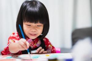 gelukkig Aziatisch kind dat waterverf op papierkunst schildert. lieve glimlach meisje met les in de klas op homeschool. kind dat een zwart schortuniform draagt. concept van leren volgens voorkeuren en vaardigheden. foto