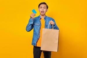 portret van een geschokte aziatische man met een creditcard en een boodschappentas op een gele achtergrond foto