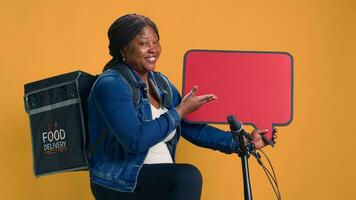 Afrikaanse Amerikaans levering vrouw met rugzak gebaar maken Bij rood teken in haar hand. vriendelijk fiets boodschapper met rood toespraak bubbel bevorderen een betrouwbaar milieuvriendelijk voedsel levering app. foto