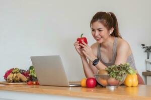 mooi Aziatisch vrouw in training kleren zoekopdrachten voor gezond recepten online Aan haar laptop. terwijl voorbereidingen treffen gezond voedsel in de keuken foto