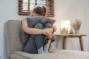 jong Aziatisch vrouw zittend met haar knieën geknuffeld, gebruik makend van een hoofdkussen naar Hoes haar gezicht Aan de sofa met spanning en ongerustheid. foto