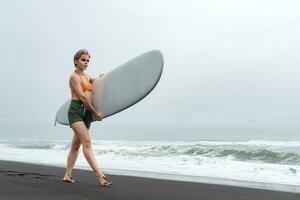 vrouw surfer wandelingen langs zwart zand strand terwijl draag- surfboard tegen backdrop van oceaan golven foto