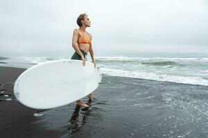 surfer staand Aan strand Holding surfboard gedurende zomer vakantie. slank vrouw op zoek Bij oceaan golven foto