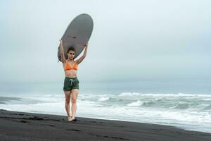 vrouw surfer wandelen Aan zwart zanderig strand met wit surfboard Aan haar hoofd gedurende zomer vakantie foto