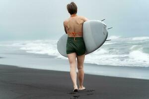 achterzijde visie onherkenbaar vrouw surfer wandelen strand draag- surfboard Aan achtergrond van zee golven foto