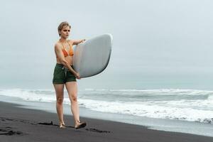 sportvrouw surfer wandelen Aan zwart zand strand, draag- wit surfboard Aan achtergrond van zee golven foto