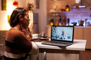 vrouw met ziekte roeping dokter Aan online video conferentie terwijl zittend in feestelijk keuken Bij huis. ziek jong geduldig gebruik makend van telegeneeskunde voor genezing behandeling en gezondheidszorg advies foto