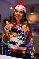 vrolijk volwassen verstrikt in Kerstmis draad lichten gebruikt voor decoreren huis. feestelijk Kaukasisch jong vrouw gebruik makend van slinger met verlichte bollen voor seizoensgebonden decor en winter boom ornamenten foto