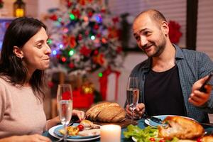 gelukkig familie aan het eten heerlijk avondeten zittend Bij dining tafel in Kerstmis versierd keuken vieren Kerstmis vakantie. paar aan het kijken vermaak film tonen gebruik makend van telefoon. Kerstman seizoen foto