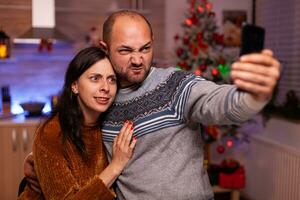 geamuseerd paar nemen selfie gebruik makend van smartphone terwijl vieren Kerstmis vakantie staand in Kerstmis versierd keuken. gelukkig blij familie genieten van uitgeven tijd samen gedurende winter seizoen foto