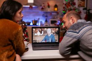 gelukkig familie pratend met afgelegen opa gedurende online video-oproep vergadering teleconferentie zittend Bij tafel in Kerstmis versierd keuken. blij paar vieren Kerstmis vakantie foto