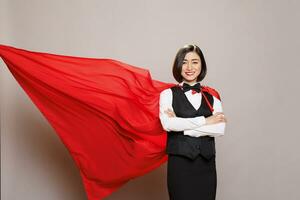 glimlachen Aziatisch serveerster vervelend superman rood mantel fladderend achter op zoek Bij camera. vrolijk vrouw receptioniste in catering onderhoud uniform en held kaap staand met armen gekruiste portret foto