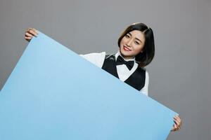 vrolijk Aziatisch vrouw receptioniste in uniform poseren met blanco blauw banier mockup en op zoek Bij camera. glimlachen serveerster Holding leeg whiteboard met copyspace portret foto