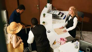 mensen aankomen Bij hotel voorkant bureau, voorbereidingen treffen naar Doen controleren in Bij ontvangst teller en zien kamer reservering. gasten Aan huwelijksreis gedurende zomertijd pratend naar conciërge in lobby. handheld schot. foto