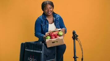 vrouw Afrikaanse Amerikaans koerier voorzichtig vervoeren fruit mand van voedsel levering tas. opgewonden levering vrouw Aan haar fiets grijpen een doos van vers en gezond produceren voor leveren. foto