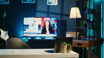 pannen schot van werk bureau met laptop en mini huis planten in vaag lit leeg huis studio interieur. appartement verlichte door rgb lichten met TV nieuws uitzending rennen in achtergrond foto