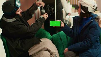 vrouw houdt smartphone met groene scherm terwijl ze praat naar vriend over winter sport- Bij berg ski toevlucht. mensen uitgerust met skiën uitrusting en bescherming, geïsoleerd mockup sjabloon. foto
