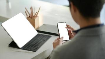 jonge zakenman houdt een smartphone met een leeg scherm in de hand en een laptop met een leeg scherm op het bureau. foto