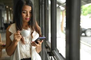 aziatische vrouw in een witte jurk die een koffiekopje in de hand houdt, gebruikt een mobiele telefoon. foto