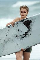 vrouw surfer draag- surfboard is wandelen Aan strand, op zoek Bij camera met zee golven in achtergrond foto