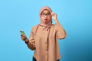 portret van een verraste jonge aziatische vrouw die een mobiele telefoon vasthoudt en een bril aanraakt met de vinger foto