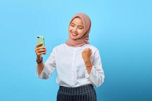 portret van vrolijke jonge aziatische vrouw houdt mobiele telefoon vast en viert succes met opgeheven hand
