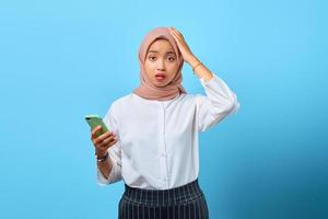portret van verraste jonge aziatische vrouw die mobiele telefoon met hand op hoofd over blauwe achtergrond gebruikt foto