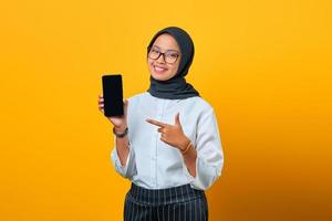 gelukkige jonge aziatische vrouw wijzend op het lege scherm van de mobiele telefoon geïsoleerd op gele achtergrond foto