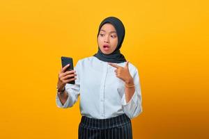 verrast jonge Aziatische vrouw wijzend op mobiele telefoon geïsoleerd op gele achtergrond