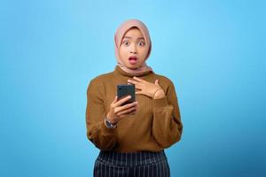 verraste jonge aziatische vrouw met mobiele telefoon met open mond op blauwe achtergrond foto