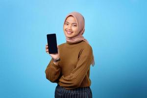gelukkige jonge aziatische vrouw die het lege scherm van de mobiele telefoon toont dat over blauwe achtergrond wordt geïsoleerd foto