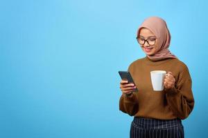 vrolijke jonge aziatische vrouw die een mok vasthoudt en op zoek is naar een smartphonescherm op een blauwe achtergrond