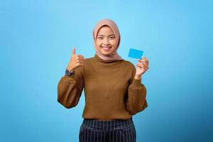 vrolijke jonge aziatische vrouw die een creditcard vasthoudt en een duim omhoog gebaar toont op een blauwe achtergrond
