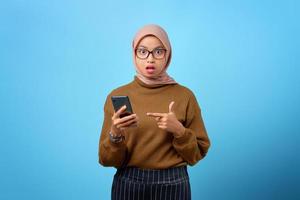 verrast jonge Aziatische vrouw wijzend op mobiele telefoon met open mond op blauwe achtergrond foto