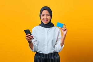 gelukkige jonge aziatische vrouw die creditcard en mobiele telefoon bij de hand op gele achtergrond toont