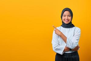 glimlachende jonge aziatische vrouw die met de vingers wijst naar kopieerruimte op gele achtergrond