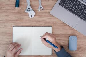 top visie foto de technicus werken Bij hout tafel en gebruikt een computer laptop naar analyseren de gebroken van auto met Vermelding gegevens in notitieboekje in onderhoud kamer concept