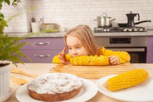 kind eet zoet gekookt maïs in Purper keuken van huis. grappig meisje Bij tafel. kronkelt. foto