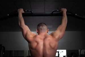 een Mens atleet bodybuilder trekt omhoog Aan zijn handen, bouwt spieren, doet een oefening in Sportschool. foto