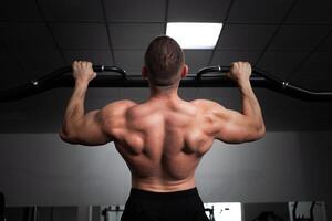 een Mens atleet bodybuilder trekt omhoog Aan zijn handen, bouwt spieren, doet een oefening in Sportschool. terug foto